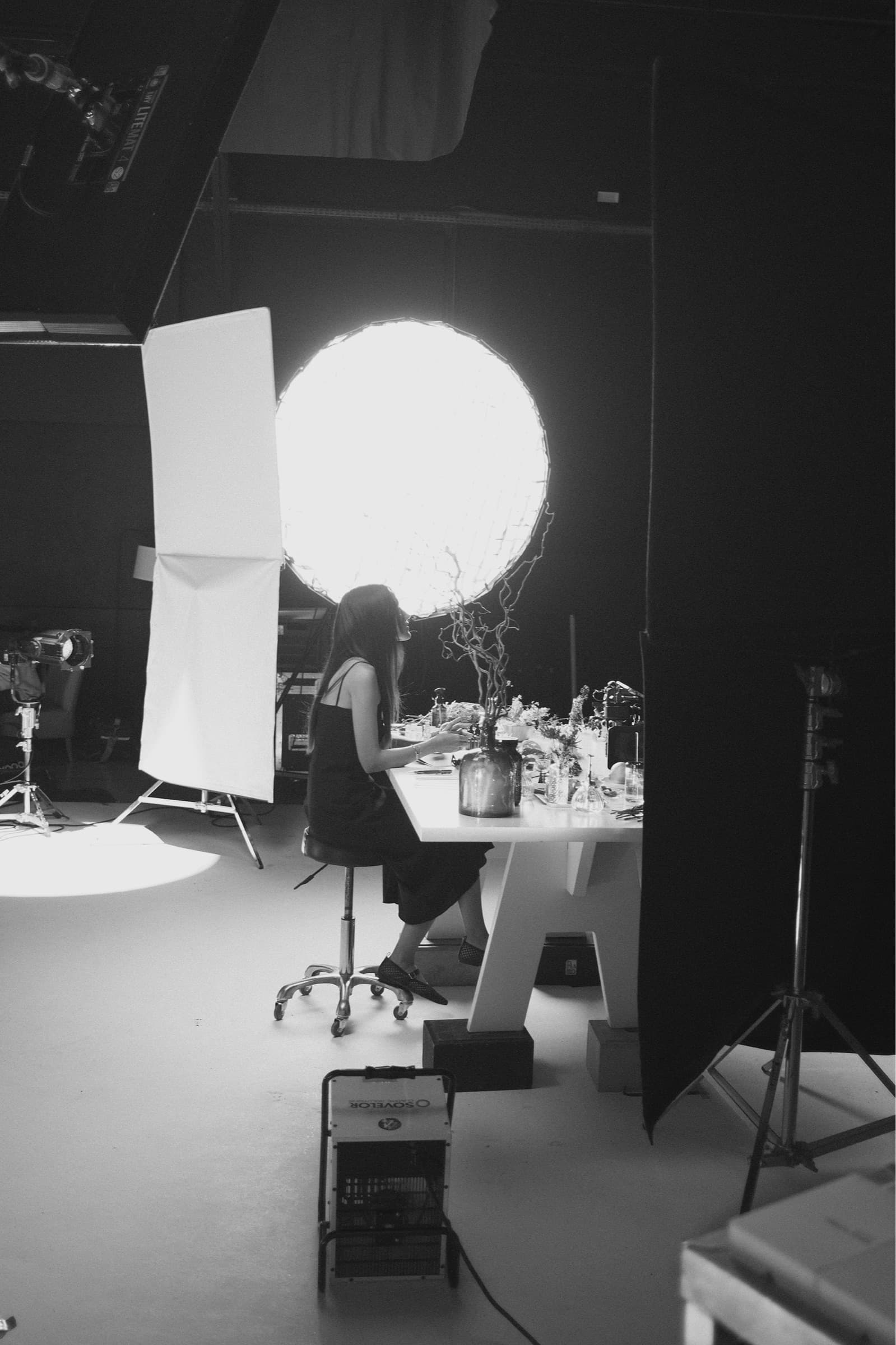 Ashley Santiago seated at a table of raw materials in a dark film studio, a large round light behind her
